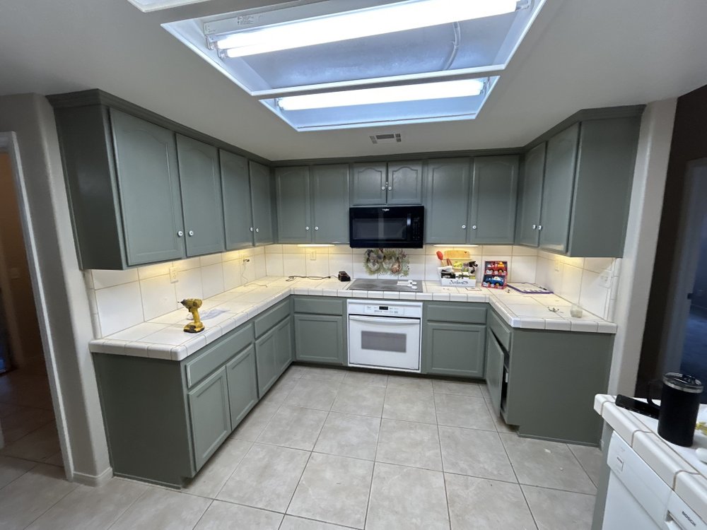 Refinished gray-green kitchen cabinets surround a microwave, dishwasher, tiled counters, and sink; a cordless drill rests on the counter. The bright kitchen has skylights, under-cabinet lighting, and a tiled floor, creating a clean, practical atmosphere.