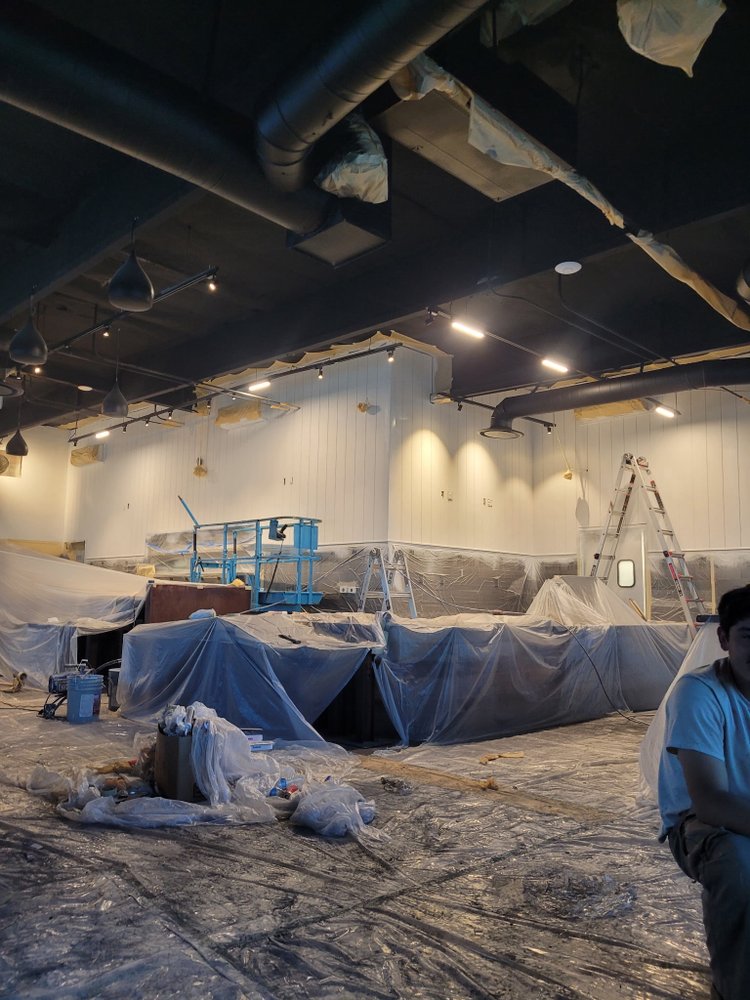 Commercial interior being prepared for painting, with exposed ceiling ducts, ladders, scaffolding, and walls partially covered in protective plastic. A worker is seated at the right edge in this busy renovation worksite.