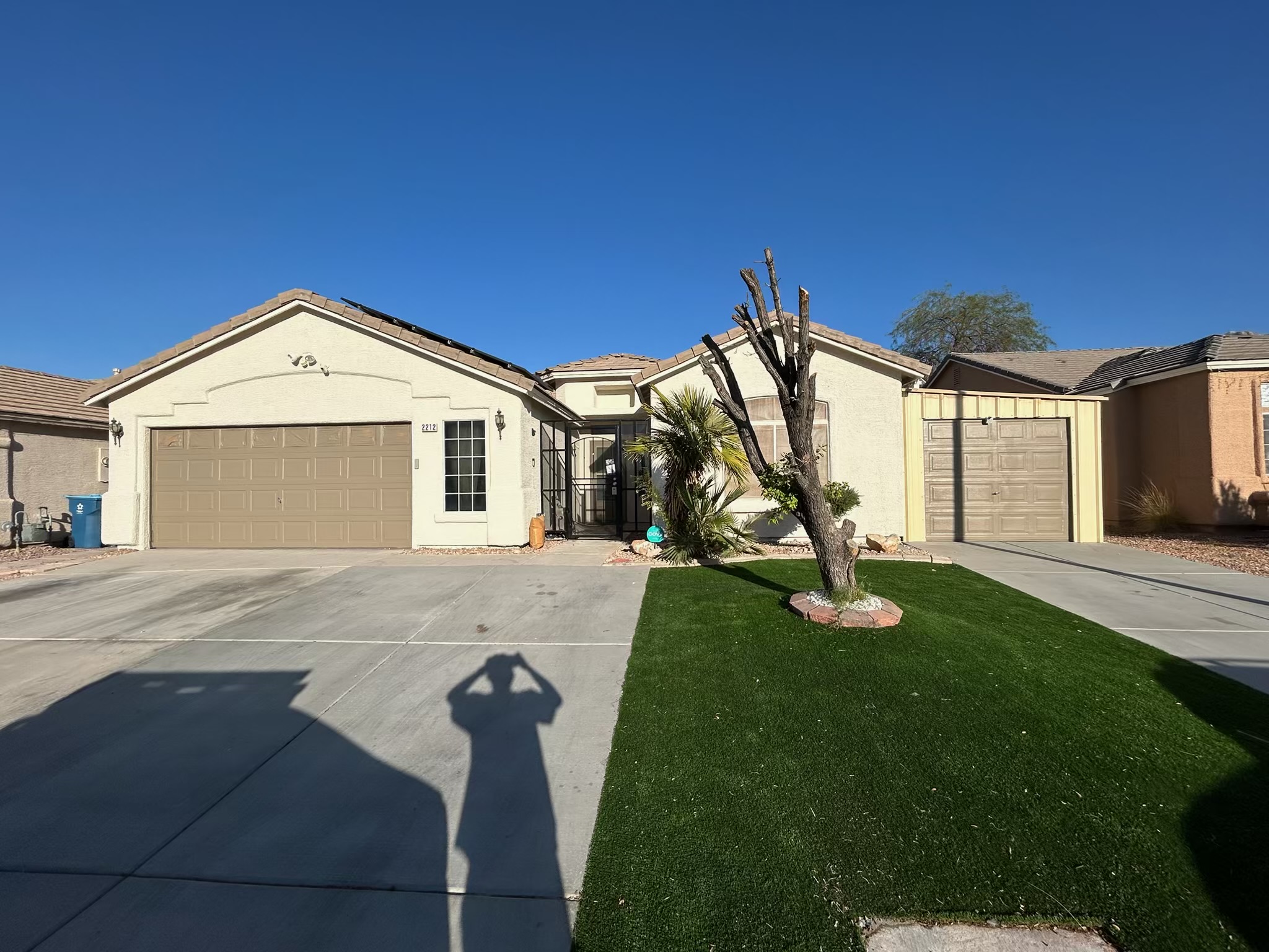 Three adjacent single-story suburban homes with pale stucco walls, tiled roofs, garages, concrete driveways, and a strip of green lawn. A pruned tree and palm plants stand in the sunny front yard beneath a clear blue sky, with the photographer shadow visible in the foreground.