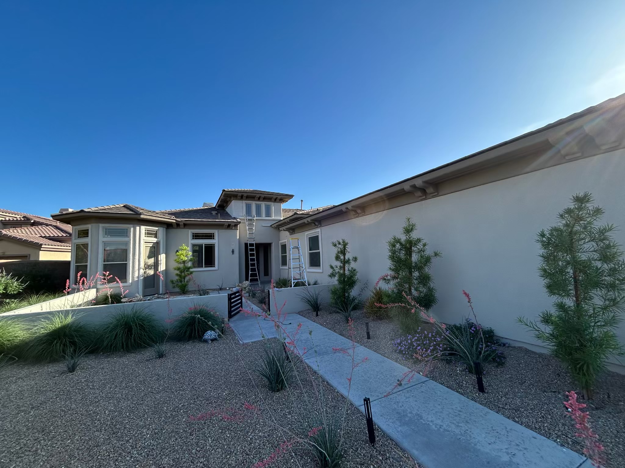 A two story tan stucco home with a light roof, a front entry, and a white accent wall. A ladder leans against the wall near the door while a concrete walkway leads through desert landscaping with grasses, shrubs, and colorful flowering plants. The sky is bright blue and the scene feels clean, calm, and sunny.