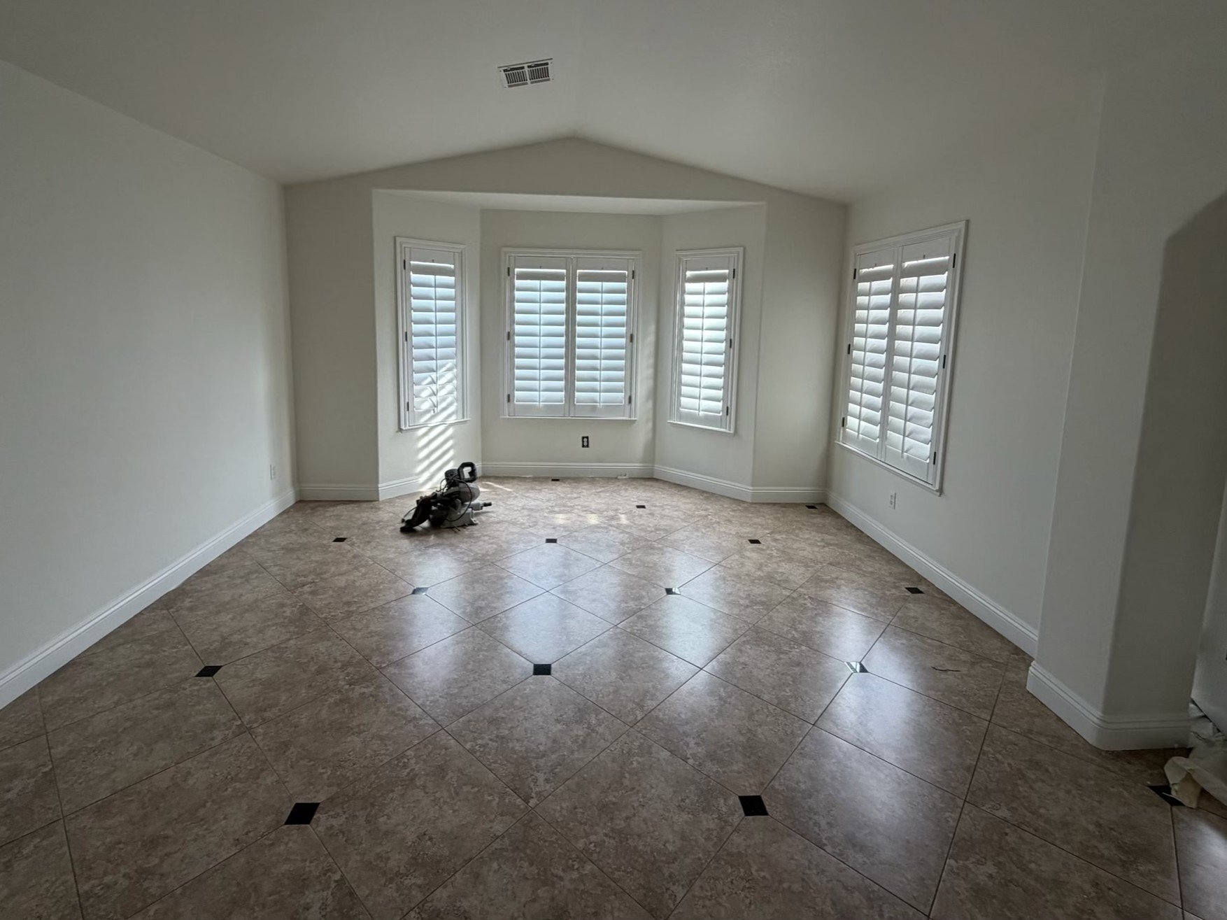 Bright, freshly painted empty room with white walls, a bay window, additional shuttered windows, white trim, and a tiled floor. Painting tools rest near the center of the room, suggesting recent interior painting work. Daylight fills the clean, quiet, and spacious room.