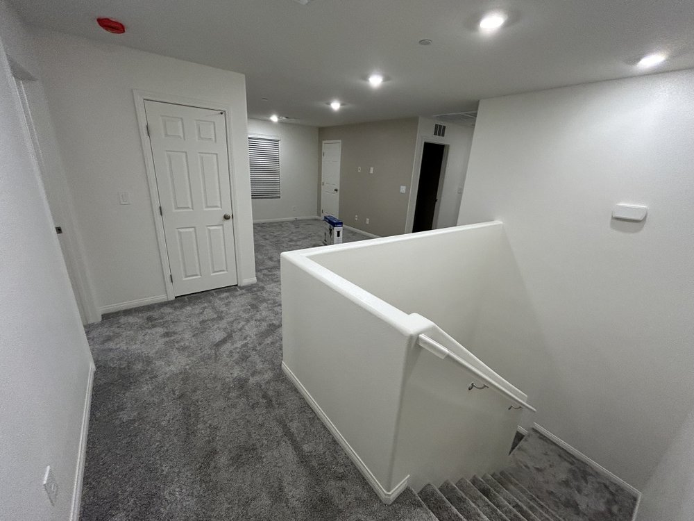 Bright, freshly painted white interior with a carpeted upstairs landing and staircase descending behind a half wall. Several closed doors, a window with blinds, recessed ceiling lights, a thermostat, and a small paint can are visible, creating a clean, newly renovated atmosphere.